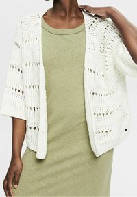 Cardigan blanco de punto abierto con mangas cortas, superpuesto a un vestido suave de color verde claro. Presenta un corte relajado y detalles de costura intrincados.
