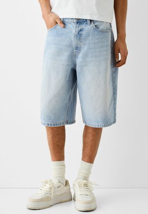 Hellblaue Jeansshorts mit lockerem Schnitt, ausgestattet mit fünf Taschen, Reißverschluss und umgeschlagenen Säumen. Kombiniert mit weißen Sneakers und Socken.