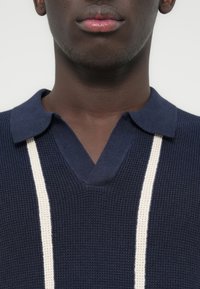 Μπλε σκούρο πλεκτό πόλο μπλουζάκι με V-λαιμόκοψη και δύο κάθετες κρεμ ρίγες στο εμπρός μέρος. Υφή υφάσματος με γιακά.