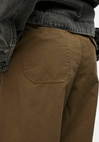 Bruine katoen broek met een grote, schuin geplaatste achterzak met een stikselrand, gecombineerd met een donkergrijze denimjack.