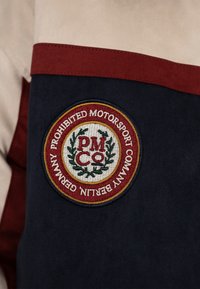 Patch rond brodé sur tissu foncé avec bordure rouge, couronne de laurier verte et texte "PM Co Prohibited Motorsport Company Berlin Germany."