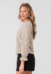 Beige bloemenkant top met lange mouwen en gerimpelde manchetten, gecombineerd met een zwarte rok. Het kant heeft ingewikkelde bloempatronen.
