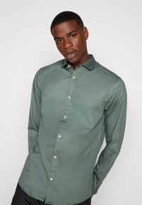 Chemise verte à manches longues avec boutons, en tissu lisse, dotée d'un col classique, avec une patte de boutonnage et une fermeture par boutons.