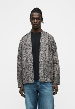 Młody mężczyzna o ciemnej karnacji i splecionych włosach, ubrany w czarną koszulę, wzorzystą szarą otwartą kurtkę oraz niebieskie dżinsy, stoi na białym tle.