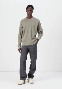 Hellgrünes, geripptes Langarmshirt kombiniert mit grauen Cargo-Hosen mit mehreren Taschen, getragen mit beigen Sneakers vor einem schlichten Hintergrund.