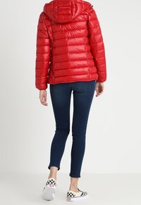 Giacca puffa rossa con cappuccio, caratterizzata da una texture trapuntata e una cerniera frontale. Indossata con jeans skinny blu scuro e scarpe slip-on a quadri.