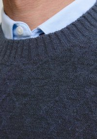 Pogled od blizu na osobu koja nosi svijetloplav izbrijani košulju ispod tamnoplave strukturirane pletene veste.