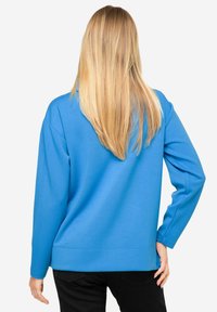 Sudadera de manga larga y cuello redondo en azul brillante, con un corte relajado, textura suave y puños acanalados. Se ven parcialmente pantalones negros.