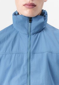 Lys blå zip-up jakke med høy krage, laget av lett stoff. Har sentral glidelås og ren sømdetalj.