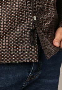 Bruine blouse met patroon en geometrische ontwerpen in oranje en blauw. Etiket genaaid aan de zoom, gedragen met blauwe spijkerbroek.