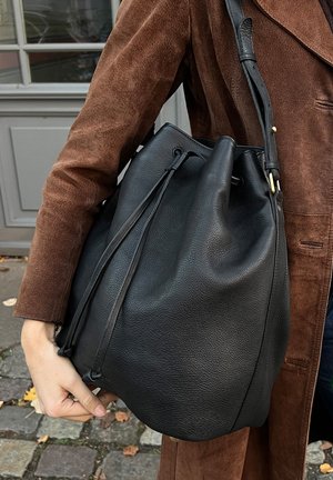 Person wearing brown suede jacket carrying large black leather bucket bag with drawstring and shoulder strap outdoors.