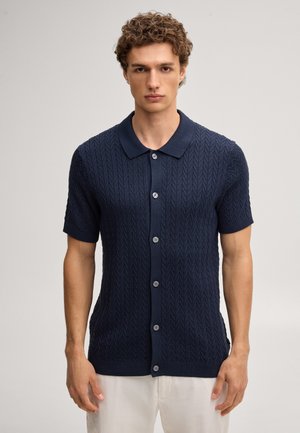 Mann mit lockigem Haar, der ein marineblaues, kurzärmeliges Strickhemd mit Kragen und weißen Hosen trägt, vor einem schlichten Hintergrund stehend.