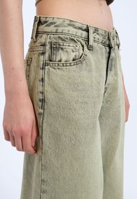 Jeans de mezclilla verde con un corte relajado, cintura alta y una sutil textura veteada. Diseño de cinco bolsillos con detalles de herrajes metálicos.
