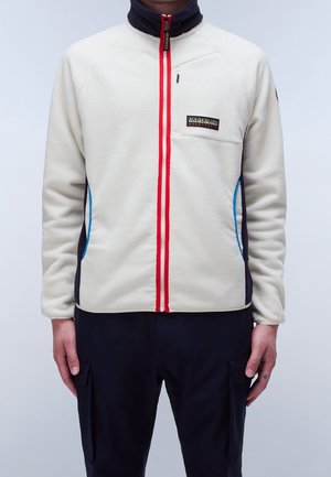 Personne portant une veste polaire couleur crème avec une fermeture éclair rouge, des accents bleus sur les côtés, une poche poitrine et un pantalon noir avec poches cargo latérales.