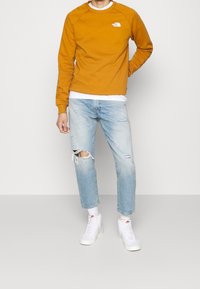 Sudadera naranja con un logo blanco, combinada con unos vaqueros rotos de color azul claro y zapatillas blancas. Ajuste casual, mangas raglán, costuras visibles.
