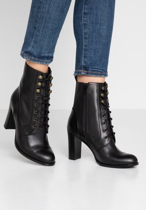 Mustat nahkaiset nilkkurit, joissa on paksut korot, etunauhat ja sivuvetoketjut. Kengät ovat yhdistetty sinisiin käännettyihin skinny-farkkuihin valkoisella lattialla.