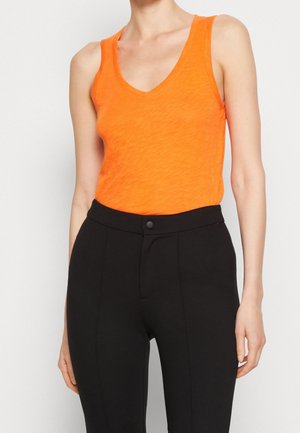 Person iført en lys orange, ærmeløs V-hals top, der er stoppet ned i højtaljede, sorte, skræddersyede bukser med en frontknap.