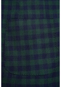 Gros plan sur un tissu à carreaux vert et bleu marine avec une poche rectangulaire cousue sur un matériau texturé.