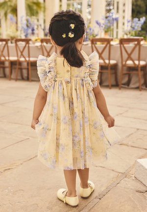 Young girl with braided hair adorned with yellow flowers, wearing a yellow floral dress and matching shoes, standing on a stone floor indoors.