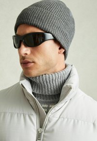 Căciulă gri cu model în riduri, ochelari de soare negri și o geacă de iarnă light grey peste un pulover gri cu guler înalt, evidențiind diverse texturi și straturi.