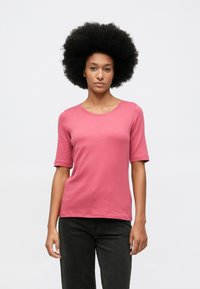 Γυναίκα με αφρο hairstyle, φορώντας ροζ κοντομάνικο μπλουζάκι και μαύρο παντελόνι, στέκεται μπροστά σε λευκό φόντο.