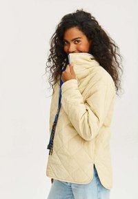 Beige prošívaná bunda s vysokým límcem, kontrastním modrým zipem a bočním rozparkem; má stahovací šňůrky s korálky pro ozdobu.