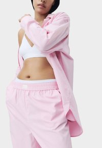 Chemise boutonnée rose clair en tissu doux, portée par-dessus un bralette blanc, assortie à un pantalon de pyjama rose avec une taille élastique.