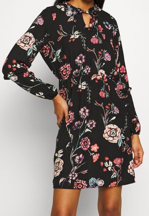 Frau trägt ein schwarzes, floral gemustertes Langarmkleid mit rosa, weißen und lila Blumen, gebundenem Ausschnitt und elastischen Manschetten, die Hand auf der Hüfte.