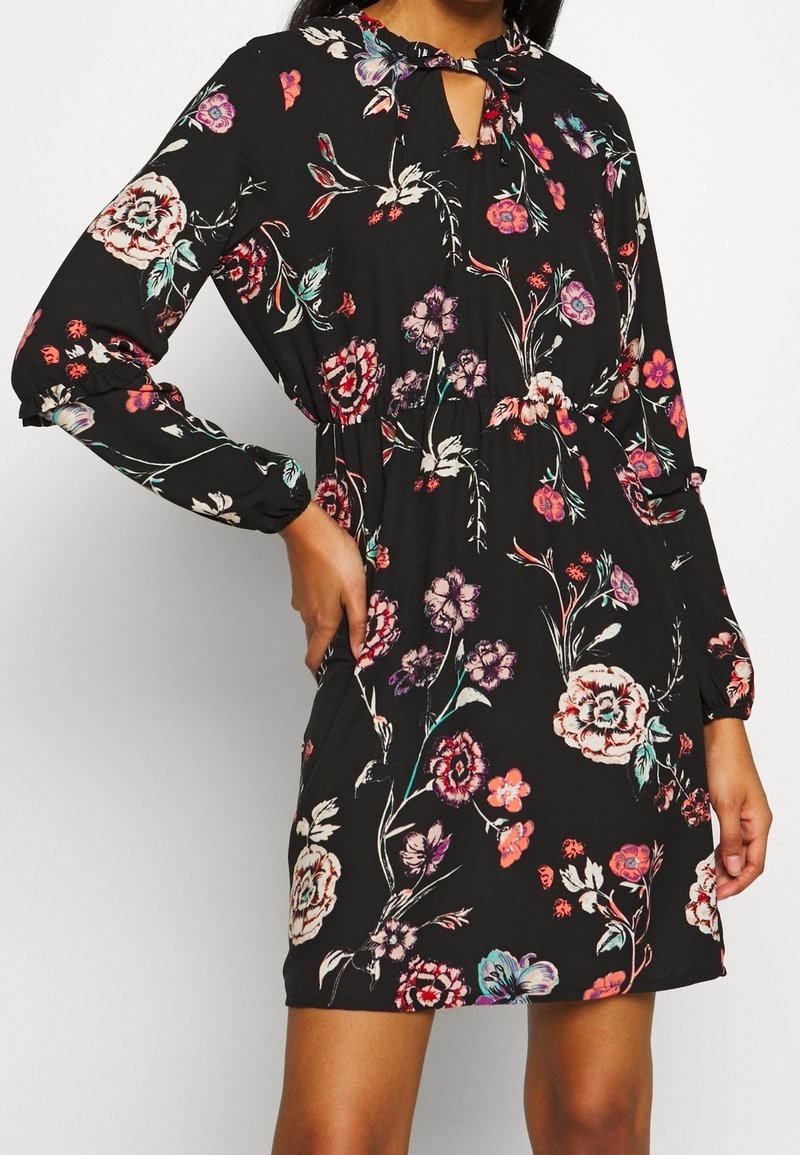 Frau trägt ein schwarzes, floral gemustertes Langarmkleid mit rosa, weißen und lila Blumen, gebundenem Ausschnitt und elastischen Manschetten, die Hand auf der Hüfte.
