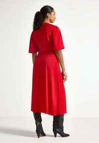 Rød knælang kjole med korte, puffede ærmer og et bælte. Modellen bærer sorte støvler i knæhøjde, der fremhæver en glat stofstruktur.