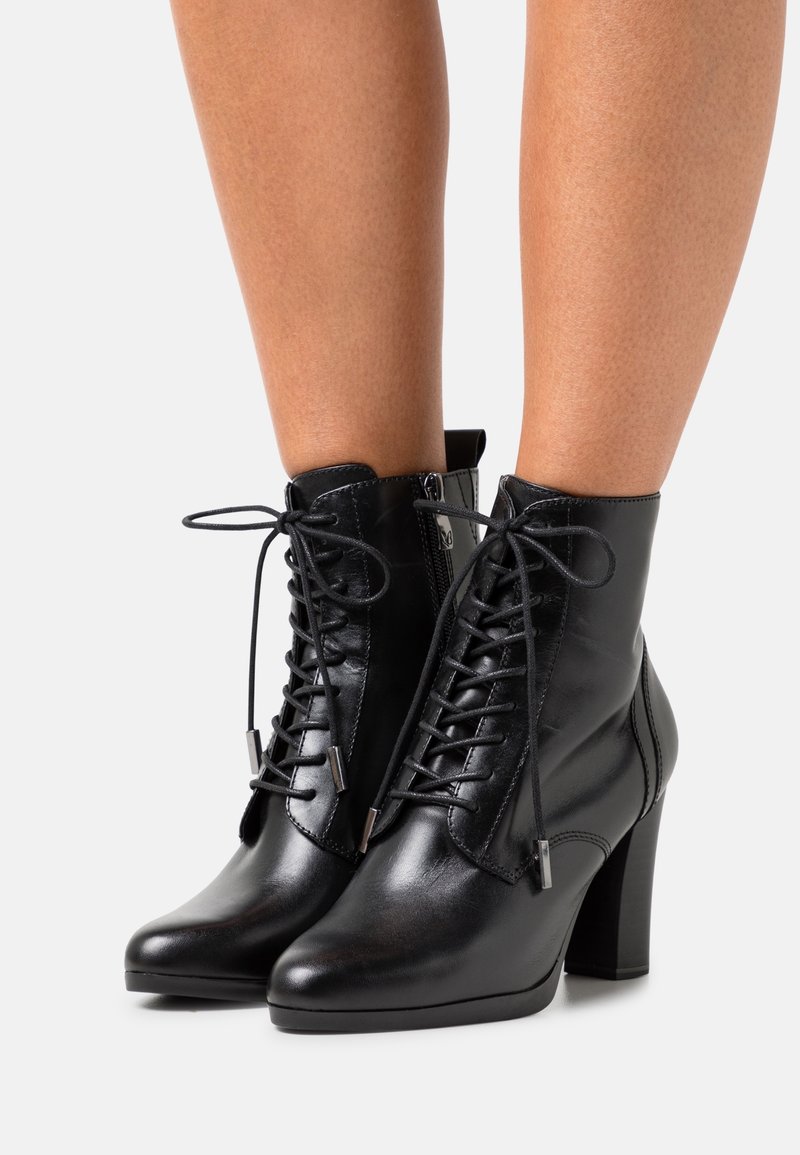 Bottines en cuir noir avec lacets et talons carrés portées sur jambes nues sur un fond uni.