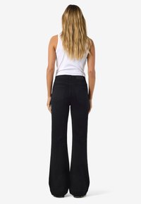 Ženska z dolgimi blond lasmi nosi belo majico brez rokavov in črne visokopasne široke hlače, stoji obrnjena stran na belem ozadju.