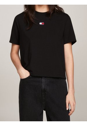 Person trägt ein schwarzes T-Shirt mit kurzen Ärmeln und kleinem Tommy Jeans-Logo auf der Brust sowie schwarze High-Waist-Jeans, eine Hand in der Tasche.