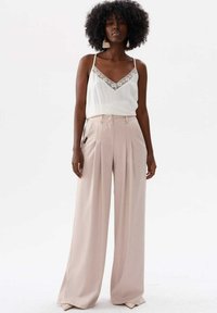 Jasna kremowa koszulka na ramiączkach z koronkowym wykończeniem, zestawiona z różowymi, szerokimi spodniami. Modelka stoi, prezentując luźny krój i gładki materiał.