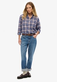 Blauw-wit geruit overhemd met opgerolde mouwen, gecombineerd met lichtblauwe jeans, een bruine gevlochten riem en donkere loafers.