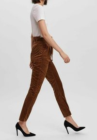 Kvinna som går iförd en vit kortärmad topp, bruna högmidjade corduroy-byxor och svarta högklackade skor mot en enfärgad bakgrund.