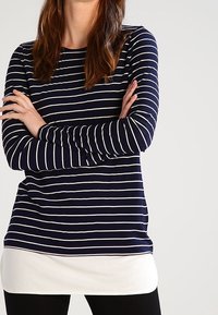 Mörkblå långärmad topp med vita horisontella ränder, med rund halsringning och lager av vit fåll, bärs över svarta leggings.