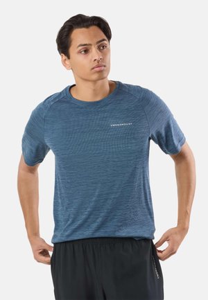 Ung mand justerer sorte shorts iført blå kortærmet sportstrøje med "Swedemount" logo på brystet mod hvid baggrund.