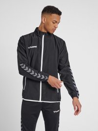Schwarze Sportjacke mit einem weißen Reißverschluss, ausgestattet mit grauen Chevron-Mustern an den Ärmeln und einem Logo auf der Brust. Glatte Textur.