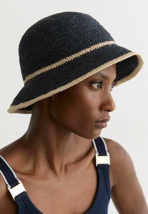 Femeie profil purtând o pălărie tip găleată din croșetat negru cu margine bej și un maiou albastru închis cu bretele cu cataramă aurie, pe un fundal simplu.