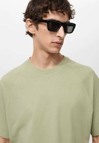 Svijetlozelena majica kratkih rukava s okruglim izrezom i raglan rukavima, uparena s crnim pravokutnim naočalama. Glatka tekstura.