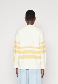Maglione in maglia crema con strisce gialle e bianche, vestibilità ampia, spalle scese e orlo a coste. Presenta un accento numerico sulla manica.