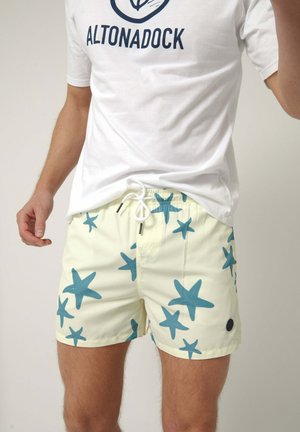 Hombre con camiseta blanca con logo y pantalones cortos de baño amarillo claro con estampados de estrellas de mar azules y cordón blanco.