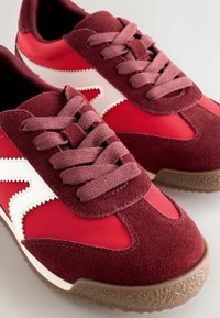 Červené sportovní boty s svrchním materiálem z semišové a syntetické kůže, zdobené bílým designovým akcentem, odpovídajícími tkaničkami a texturovanou gumovou podrážkou.