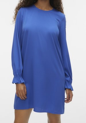 Mujer con un vestido azul real suelto, de manga larga y largo hasta la rodilla, con puños fruncidos, de pie contra un fondo claro y sencillo.