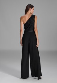 Schwarzer Jumpsuit mit einem transparenten, verzierten Ein-Schulter-Design, weit geschnittenen Hosenbeinen und einer taillierten Taille. Glatte Textur über das gesamte Kleidungsstück.
