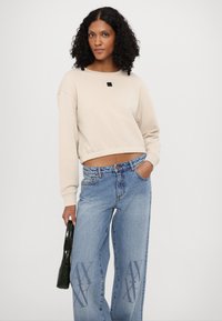 Felpa beige cropped con orlo a coste, collo rotondo e dettaglio patch nero. Abbinata a jeans a gamba larga in denim blu con vita alta e accenti con logo.
