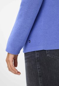 Pull en tricot violet clair avec des poignets et un ourlet côtelés. Présente un petit logo rond sur le côté, associé à un jean en denim foncé.