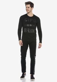 Schwarzes Langarmshirt mit erhabenen Textgrafiken mit "CIPO" und "DENIM BRAND." Kombiniert mit schwarzen Hosen, die einen Seitenaufdruck zeigen.