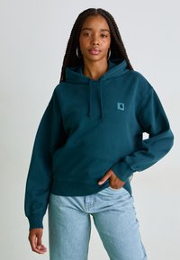 Sweatshirt à capuche teal en coton, avec une poche avant, des poignets côtelés, et un petit patch logo sur la poitrine, associé à un jean bleu clair.